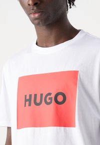 Camiseta de algodón blanca con un gran logotipo rectangular rojo que lleva la palabra "HUGO" en letras negras en negrita y mayúsculas. Diseño clásico de cuello redondo.