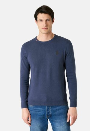 Uomo che indossa un maglione blu navy a maniche lunghe semplice e jeans blu, in piedi contro uno sfondo bianco.