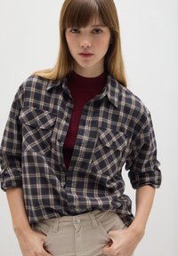 Camisa de franela de cuadro en azul oscuro y beige, con dos bolsillos en el pecho y cierre de botones en la parte frontal. El modelo lleva un jersey de cuello alto burdeos debajo.