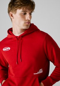 Rote Kapuzenjacke aus weichem Material mit einer Kängurutasche, versehen mit einem weißen Logo auf der Brust und dem Schriftzug „RED BULL RACING WORLDWIDE.“