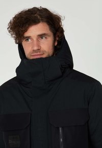 Svart vattentät jacka med hög krage, framdragkedja, två stora fickor med klaff och texturerade detaljer på axlarna.