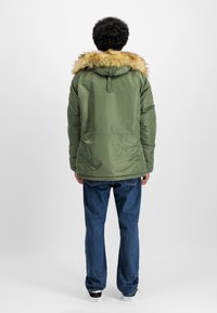 Green insulated jacket with a faux fur hood, elastic cuffs, and a smooth finish. Paired with blue denim jeans and white sneakers.