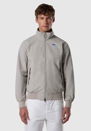 Junger Mann mit kurzen braunen Haaren trägt eine hellgraue Jacke mit Reißverschluss und einem runden Kompass-Logo auf der Brust sowie weiße Hose, steht vor einem einfarbigen Hintergrund.