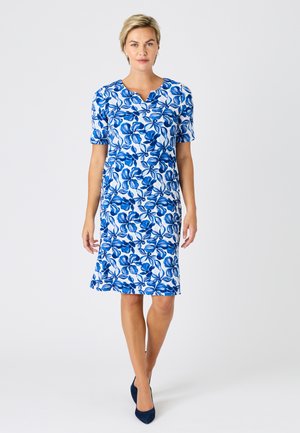 Femme debout portant une robe blanche mi-longue à motifs floraux bleus et des talons bleu marine, sur un fond blanc uni.