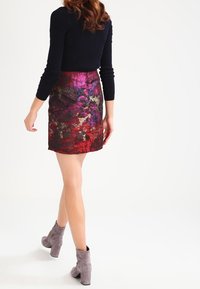 Femme marchant avec un haut noir à manches longues, une jupe à motifs abstraits multicolores et des bottines en daim grises contre un fond blanc.