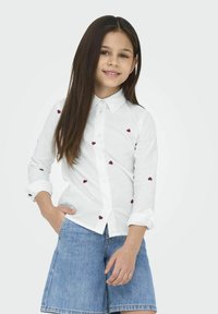 Camisa blanca de botones con corazones rojos bordados, mangas largas enrolladas en los puños, combinada con pantalones cortos de mezclilla azules. Textura de tela suave.