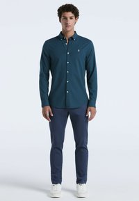 Teal overhemd met knopen en een subtiele textuur, met een klein wit logo. Gecombineerd met navy broek en witte sneakers.