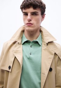 Jeune homme aux cheveux courts et bouclés portant un trench-coat beige sur un polo vert clair, regardant directement la caméra avec une expression neutre.