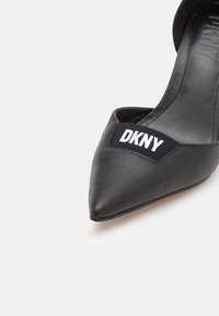 Chaussure à talon haut noire à bout pointu avec un patch logo DKNY sur la partie supérieure avant.