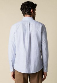 Muž oblečený v dlouhém světle modrém a bílém tenkém proužkovaném tričku s dlouhým rukávem a hnědých kalhotách, viděný zezadu na neutrálním pozadí.