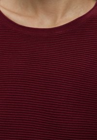 Gros plan sur le cou et le haut de la poitrine d'une personne portant une chemise texturée, côtelée, rouge foncé ou bordeaux.