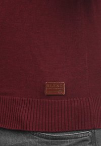 Burgunderfarbener Strickpullover mit geripptem Saum und einem braunen Lederetikett mit der Aufschrift "BLEND", das an der Vorderseite nahe dem unteren Rand angenäht ist.