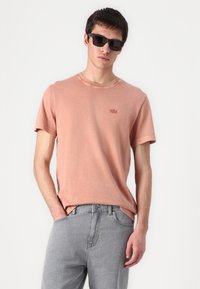 Tricou din bumbac roz deschis, cu mâneci scurte, guler rotund și un mic logo brodat pe piept, asortat cu blugi gri din denim.