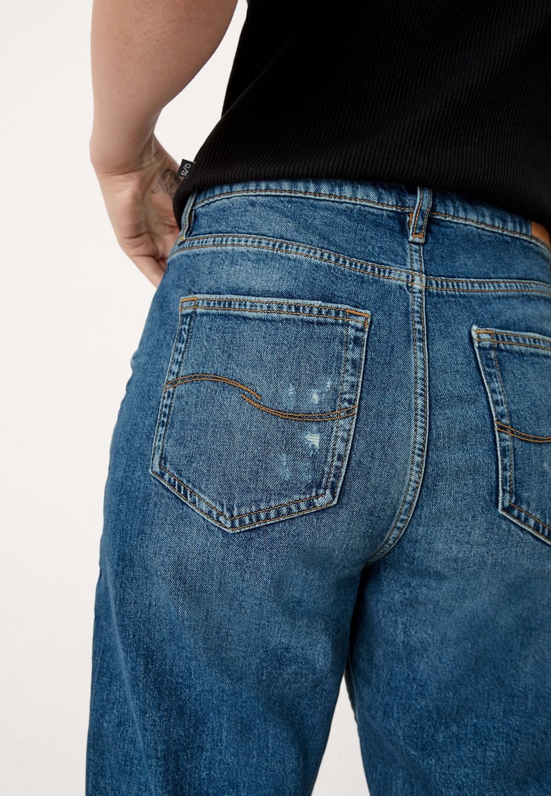 Gros plan d'une personne portant un jean bleu en denim avec des poches arrière visibles et des marques d'usure subtiles sur la poche gauche, associé à un haut noir.
