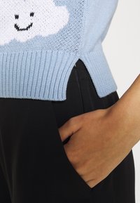 Jasnoniebieski sweter z dzianiny z białą chmurką i uśmiechniętą twarzą, prążkowany dół, boczne rozcięcia, w połączeniu z czarnymi spodniami.