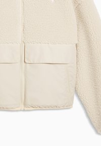 Μπεζ μπουφάν sherpa fleece με φερμουάρ μπροστά και δύο μεγάλες τσεπές με καπάκι στο κάτω μέρος, που δείχνει αντίθεση υφής μεταξύ των υλικών.