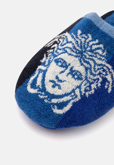 Gros plan d'une pantoufle en tissu bleu présentant un visage brodé en blanc et bleu foncé avec un motif de cheveux fluides.