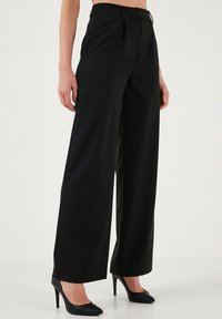 Black wide-leg trousers crafted from smooth fabric, featuring pleats, a button closure, and a high waist. Worn with black pointed heels.