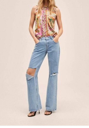 Kvinde iført ærmeløs farverig mønstret top, lyseblå revnede vide jeans og sorte sandaler med hæl og stropper.