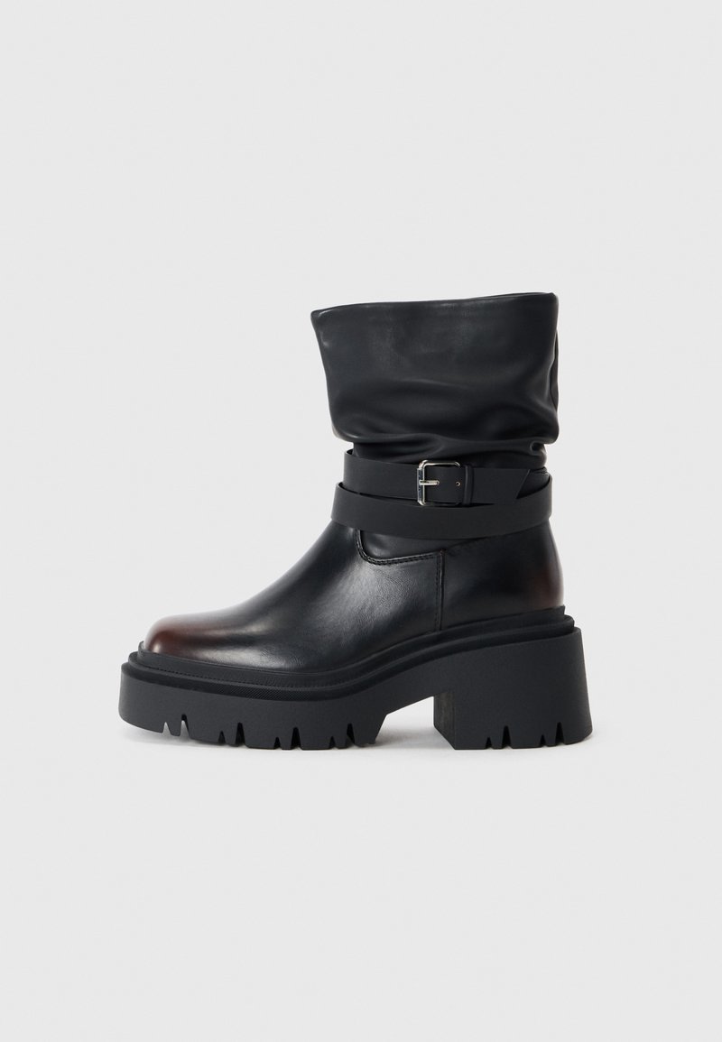 Sort læderankelstøvle med en slouchy overdel, spænde detaljer og en tyk, robust sål. Har rund tå og glat tekstur.