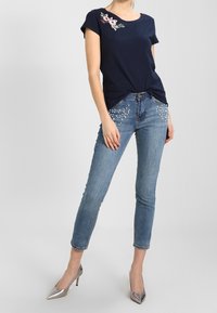 Mujer vestida con una blusa de manga corta azul marino con bordado floral, jeans cortos azul claro con detalles de perlas y tacones plateados de punta.