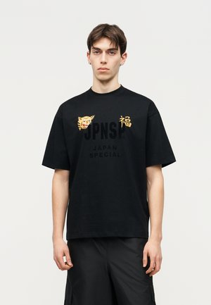 Junger Mann trägt ein schwarzes übergroßes T-Shirt mit Tigerkopf- und goldenen japanischen Schriftzeichen, kombiniert mit schwarzen Shorts, neutraler Hintergrund.