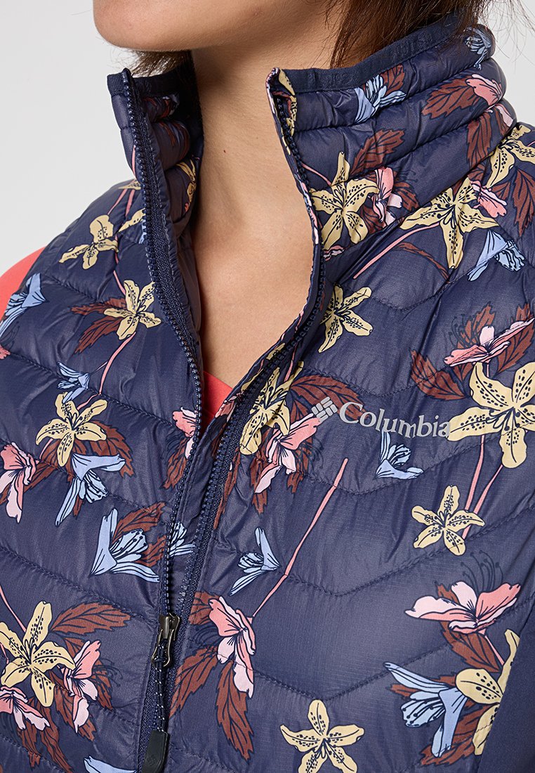 Chaleco de plumas floral azul marino con cuello alto, adornado con flores en tonos rosa claro y amarillo. Cierre de cremallera y logo de Columbia en el pecho.