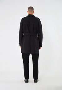 Svart trench coat med bälte i midjan, långa ärmar och axelpartier, i kombination med svarta byxor och skor, sedd bakifrån.