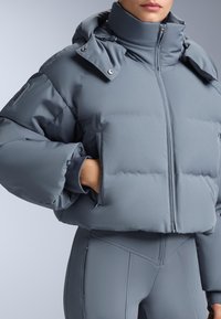 Siva skraćena puf jakna s visokim ovratnikom, zatvaranjem na patentni zatvarač i bočnim džepovima. Glatki, fleksibilni materijal s mat završnom obradom.