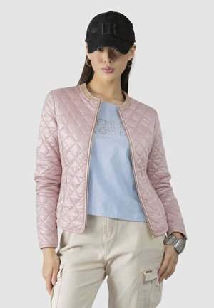 Donna che indossa una giacca trapuntata rosa chiaro, una t-shirt grafica blu, pantaloni cargo beige, un berretto nero, orecchini a cerchio e un grande bracciale d'argento.