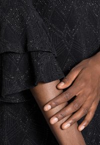 Tela de vestido negro con un patrón de diamante texturizado y acentos brillantes, que presenta volantes en las mangas y un primer plano de una mano sobre un brazo.