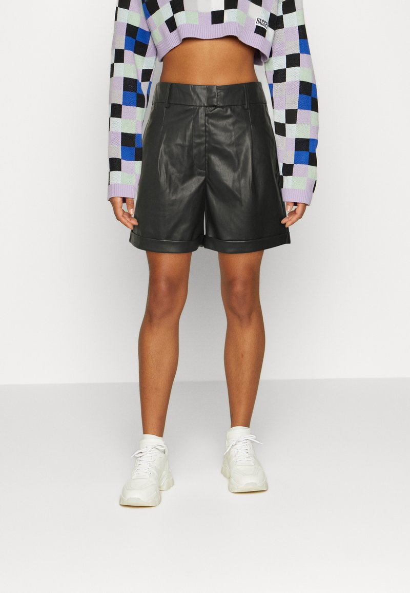 Personne portant un short en cuir noir, des baskets blanches et un pull court à carreaux pastels, vue du milieu du torse jusqu’aux pieds.