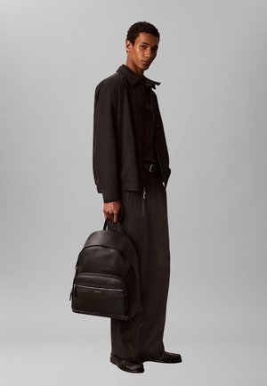 Uomo che indossa giacca e pantaloni neri, tenendo uno zaino grande in pelle nera testurizzata per la maniglia, in piedi davanti a uno sfondo grigio semplice.