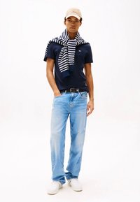 Tamsiai mėlyna polo marškinėliai, šviesiai mėlyni, nusidėvėję džinsai, balti sportbačiai ir dryžuotas tamsiai mėlynas ir baltas megztinis užmestas ant pečių. Smėlio spalvos kepurė.