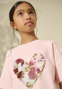 Rosa Baumwoll-T-Shirt mit einem Herzdesign aus Pailletten in verschiedenen Rosa- und Goldtönen, mit einer strukturierten, glänzenden Oberfläche.