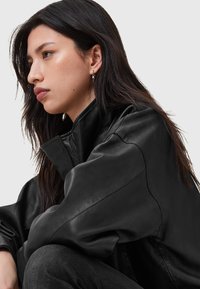 Zwarte leren jas met een hoge kraag, oversized ontwerp en een gladde textuur. Model heeft donker haar en draagt zilveren hannibal oorbellen.