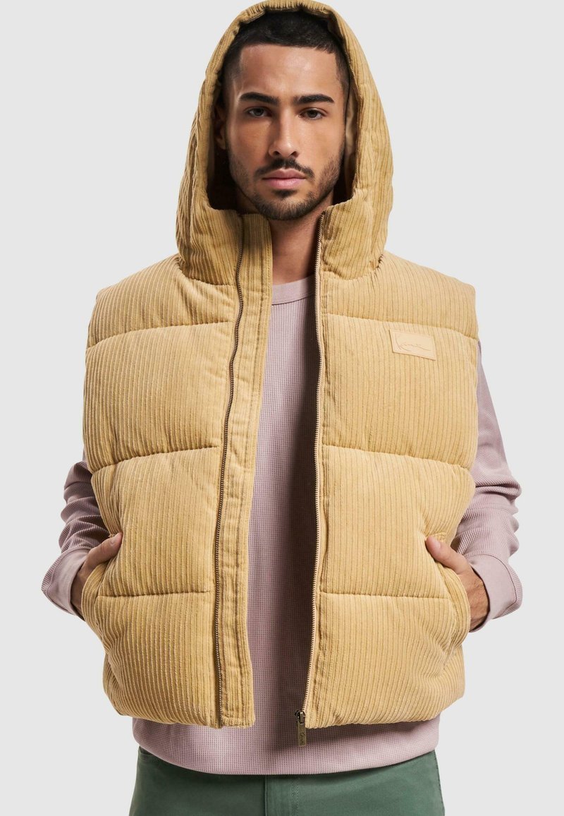 Chaleco acolchado en un cálido color beige de pana con capucha, que presenta un cierre de cremallera en la parte frontal y bolsillos laterales, superpuesto a una camiseta de manga larga en un suave color rosa.