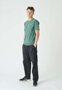 Grünes Baumwoll-T-Shirt mit kleinem blauen Logo, kombiniert mit lockeren schwarzen Hosen und schwarzen Sneakern, vor einem schlichten Hintergrund stehend.