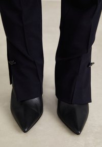 Schwarze Leder-Pumps mit spitzem Zehenabschluss, kombiniert mit taillierten schwarzen Hosen. Die Hose hat eine schmale Passform und ein dezent gearbeitetes Detail am Saum.