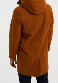 Ruskea vedenpitävä parka hupulla. Suoraviivainen leikkaus, pitkät hihat ja takana liikkuvuutta parantava halkio. Sileä, mattapintainen kangas.