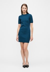 Γυναίκα που φοράει εφαρμοστό τιρκουάζ πλεκτό φόρεμα με κοντά μανίκια και σχέδια σε σχήμα ζιγκ-ζαγκ, μαζί με μαύρα παπούτσια με μυτερή μυτη, στέκεται μπροστά σε λευκό φόντο.