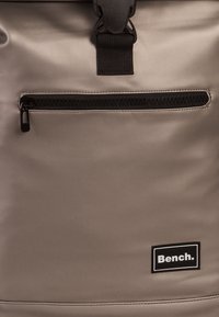 Sac à dos gris résistant à l'eau en matériau lisse, doté d'une poche zippée à l'avant, d'une fermeture à sangle noire et d'un patch logo noir.