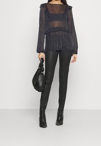 Top nero trasparente con glitter, dettagli arruffati e maniche lunghe, abbinato a pantaloni in pelle nera aderenti e una borsa nera annodata.