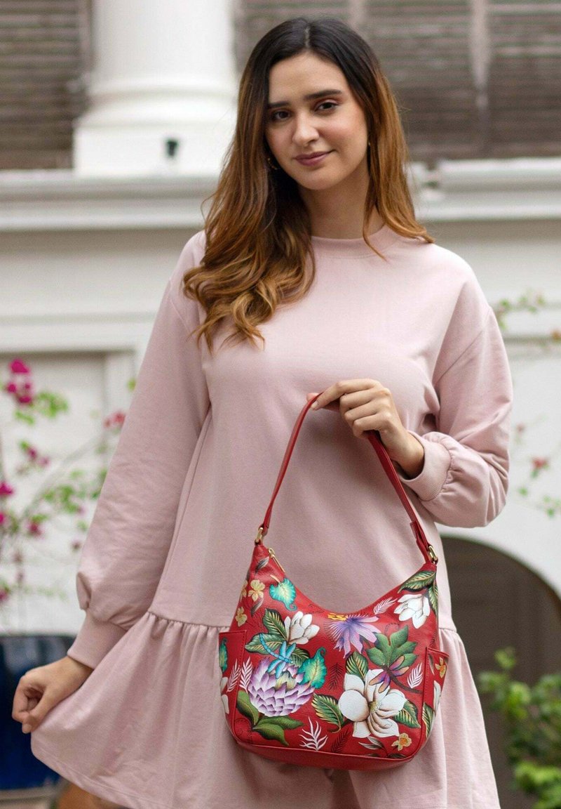 Rode leren handtas met kleurrijke bloemmotieven, zachte ronde vorm en een korte schouderband. Model draagt een lichtroze lange mouwen jurk.