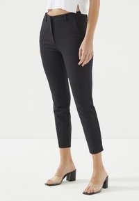 Zwarte getailleerde broek met een slim fit en cropped lengte. Glad stof met zijzakken. Gecombineerd met duidelijke hakken mules.