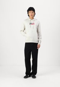 Haut en coton gris clair avec un logo sur le devant en rouge et bleu marine, doté d'une capuche avec cordon de serrage et d'une poche kangourou, associé à un pantalon noir.