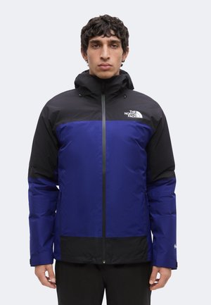 Mężczyzna w niebiesko-czarnej kurtce The North Face z kapturem, stojący zwrócony przodem na jednolitym jasnym tle.