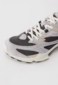 Szare i czarne sportowe buty z białymi sznurowadłami i beżowymi akcentami na białym tle, pokazujące szczegóły z przodu i z boku.