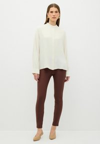 Crème-kleurige blouse met lange mouwen en een Mandarin-kraag, gecombineerd met bruine leggings en beige puntige schoenen, staande tegen een witte achtergrond.