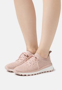 Pieds portant des baskets tricotées roses pâles avec des lacets et des semelles blanches, posés sur une surface blanche.